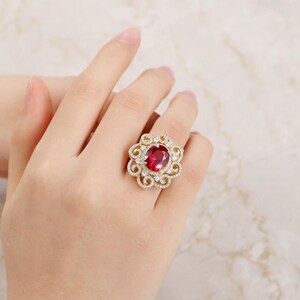 May include: A gold ring featuring a large oval red gemstone surrounded by smaller white gemstones. The ring has a delicate, floral design.