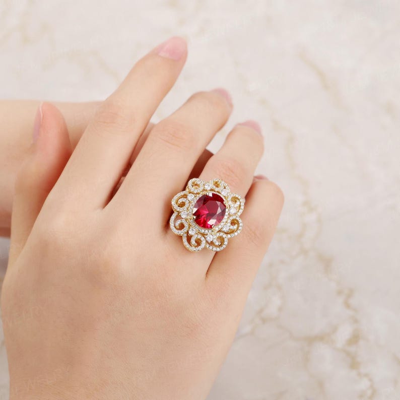 May include: A gold ring featuring a large oval red gemstone surrounded by smaller white gemstones. The ring has a delicate, floral design.