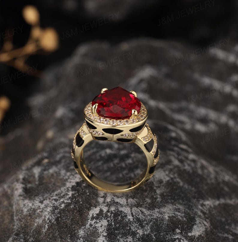 May include: A gold ring featuring a large red heart-shaped gemstone encircled by smaller clear gemstones. The band of the ring is black with a white pattern.