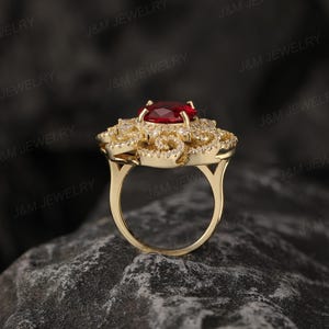 May include: A gold ring featuring a large red gemstone surrounded by smaller white gemstones. The ring has a floral design.