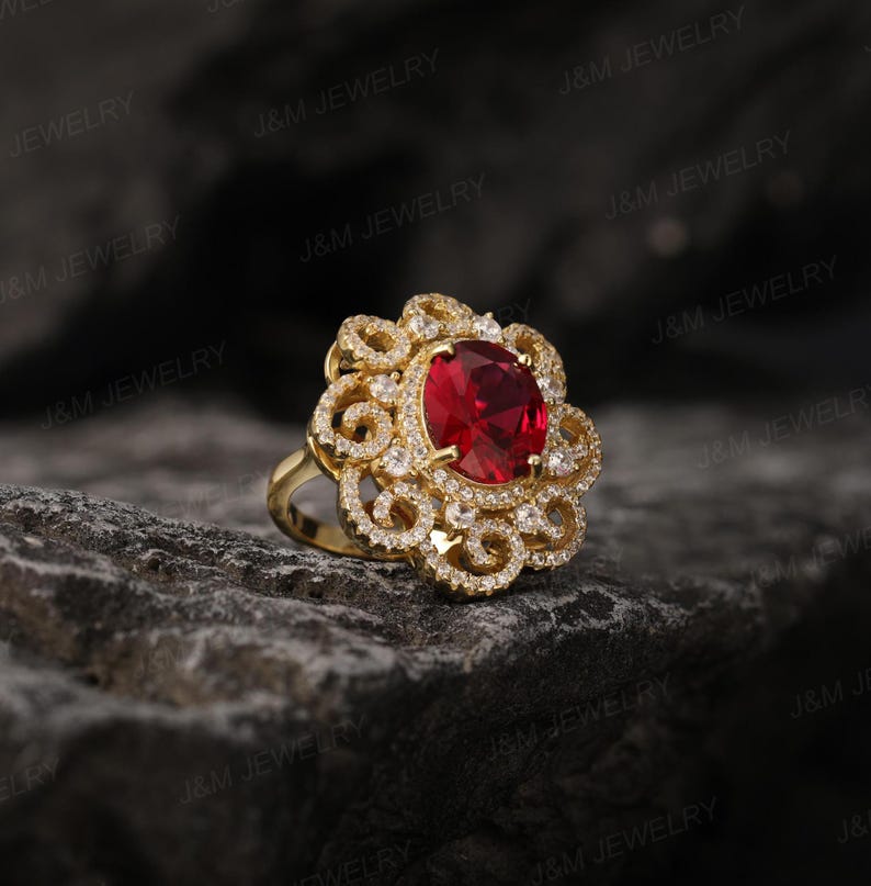 May include: A gold ring featuring a large red gemstone surrounded by smaller clear gemstones. The ring has a delicate, intricate design.