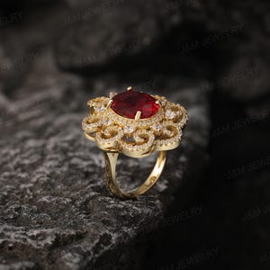 May include: A gold ring featuring a large red gemstone surrounded by smaller white gemstones. The ring has a delicate, intricate design.