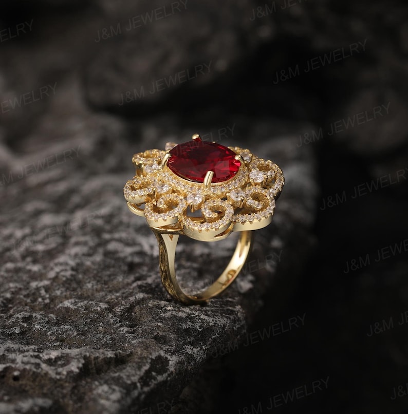 May include: A gold ring featuring a large red gemstone surrounded by smaller white gemstones. The ring has a delicate, intricate design.