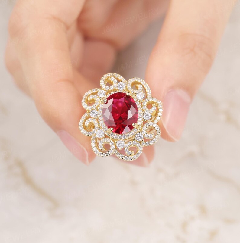 May include: A gold ring featuring a large oval red gemstone surrounded by smaller white gemstones in a floral design.