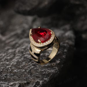 May include: A gold ring featuring a large red pear-shaped gemstone surrounded by smaller clear gemstones. The ring band is black and white.