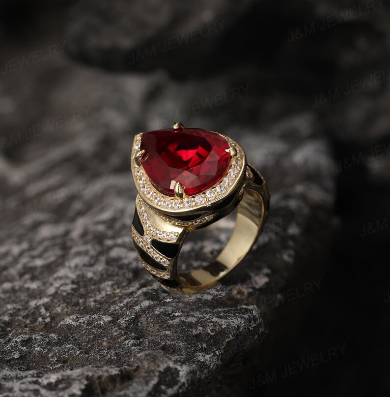 May include: A gold ring featuring a large red pear-shaped gemstone surrounded by smaller clear gemstones. The ring band is black and white.