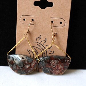 May include: A pair of dangle earrings featuring semi-circular, patterned stones in shades of brown, red, and gray. The stones are framed with gold-colored metal and suspended from gold-colored ear wires. The earrings hang from a brown card.