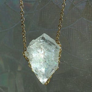 May include: A clear, rough-cut crystal pendant necklace. The crystal is suspended from a delicate gold-colored chain. The crystal has a multifaceted shape and appears to be translucent, with subtle iridescent flashes.