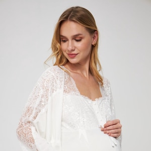 Puede incluir: Conjunto de camisón y bata de maternidad blancos. El camisón tiene un escote cuadrado y detalles de botones. La bata presenta mangas y ribetes de encaje. El conjunto está hecho de una tela suave y fluida.