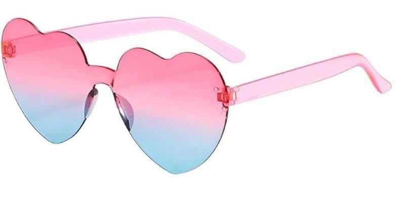 May include: Heart-shaped sunglasses featuring a rimless design with a gradient color transition from pink at the top to light blue at the bottom. The temples are made of translucent pink plastic, and the lenses are connected by a central bridge.