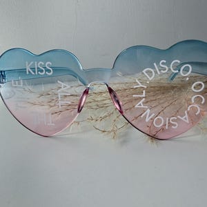 May include: Heart-shaped sunglasses with a blue-to-pink gradient tint. The left lens features the white text KISS ALL THE TIME, while the right lens displays the white text DISCO OCCASIONALLY. The frames are rimless with a bridge and nose pads.