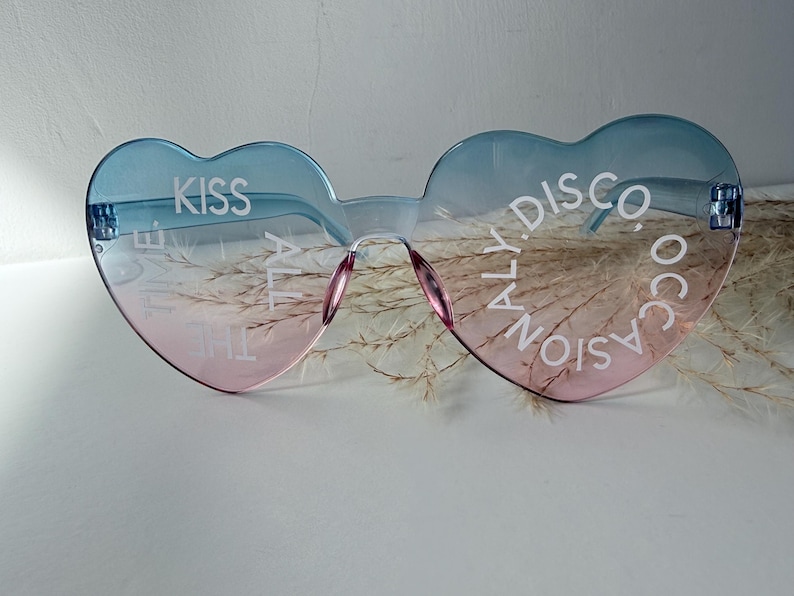 May include: Heart-shaped sunglasses with a blue-to-pink gradient tint. The left lens features the white text KISS ALL THE TIME, while the right lens displays the white text DISCO OCCASIONALLY. The frames are rimless with a bridge and nose pads.