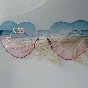 May include: Heart-shaped sunglasses with a blue to pink gradient tint. The lenses feature iridescent text that reads KISS TIME on the left and THE OCCASIONAL DISCO on the right. The frames are rimless with a thin bridge and integrated nose pads.
