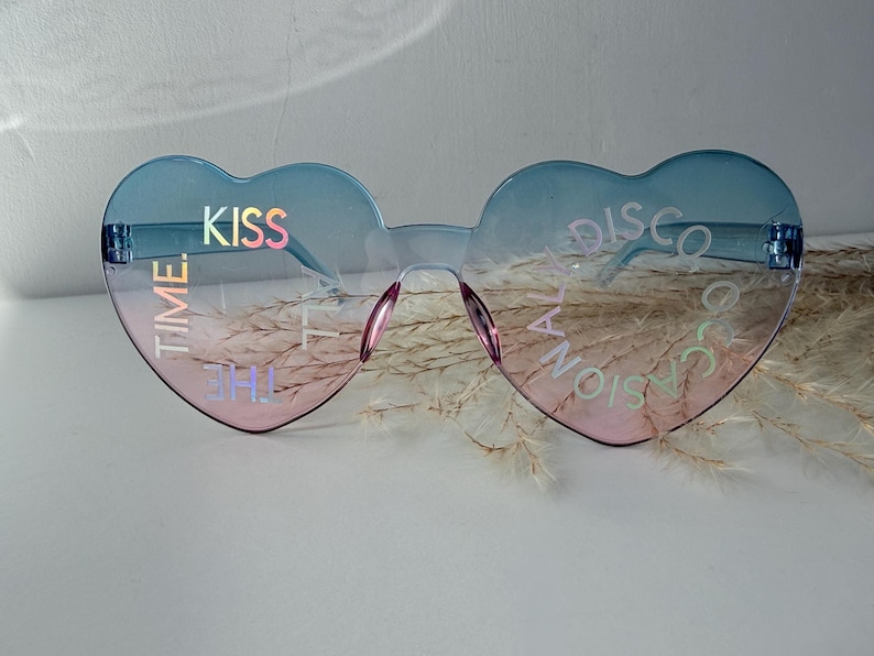May include: Heart-shaped sunglasses with a blue to pink gradient tint. The lenses feature iridescent text that reads KISS TIME on the left and THE OCCASIONAL DISCO on the right. The frames are rimless with a thin bridge and integrated nose pads.