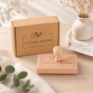 Pode incluir: Um carimbo de madeira com o texto "NATURAL BLOOM BOUTIQUE" e um design floral, juntamente com uma caixa de cart&atilde;o a condizer. A pega do carimbo &eacute; de madeira clara. Um pequeno vaso com flores secas e um cart&atilde;o de "obrigado" est&atilde;o no fundo.