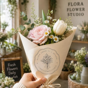 Pode incluir: Um buqu&ecirc; de flores frescas embrulhado em papel castanho. O buqu&ecirc; inclui uma rosa rosa, uma tulipa branca e pequenas margaridas brancas. O papel &eacute; impresso com o texto "FLORA FLOWER STUDIO" e "BLOOM, GREENERY & NATURAL".