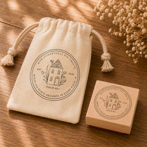 Pode incluir: Uma bolsa bege com cord&atilde;o e um carimbo de madeira com um desenho de casa e o texto "WHOLEHEARTEDLY HIS CRAFTS" e "PSALM 46:1". A bolsa e o carimbo est&atilde;o sobre uma superf&iacute;cie de madeira.