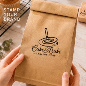 Pode incluir: Um saco de papel castanho com um log&oacute;tipo preto que diz "Cake & Bake" e um slogan. O log&oacute;tipo apresenta um batedor sobre um redemoinho. As palavras "Stamp Your Brand" est&atilde;o impressas a branco no canto superior esquerdo.