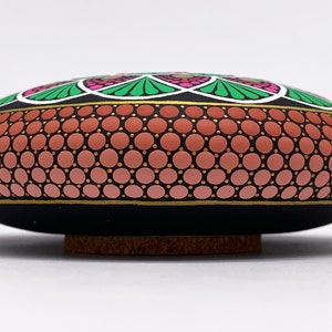 May include: A hand-painted stone featuring a mandala design. The stone is black with a pink, green and white mandala design. The stone is sitting on a wooden base.