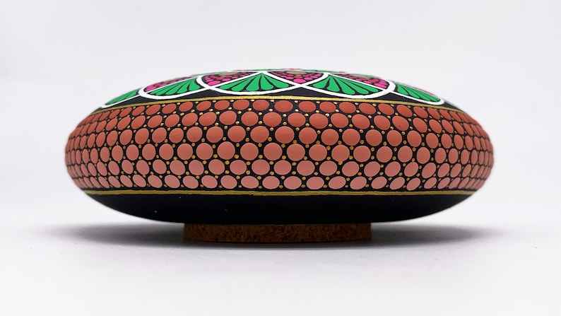 May include: A hand-painted stone featuring a mandala design. The stone is black with a pink, green and white mandala design. The stone is sitting on a wooden base.
