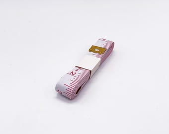 May include: A white tape measure with pink markings and a gold metal end cap. The tape measure is rolled up and the numbers 1 and 2 are visible.