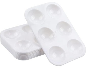 May include: A stack of white plastic paint palettes with six round wells. The palettes are designed for holding and mixing paint.