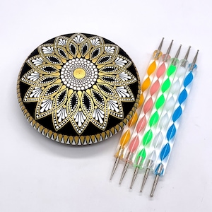May include: A black, gold, and white mandala stone with a floral design. Next to it are five dotting tools with colorful, clear handles. The mandala stone is round and features a detailed pattern with gold accents.