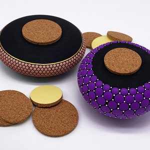 May include: Two round wooden boxes with cork lids. The box on the left is painted pink with black and gold accents. The box on the right is painted purple with black and white accents. Both boxes are decorated with a pattern of small circles.