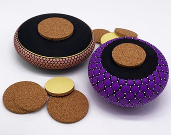 May include: Two round wooden boxes with cork lids. The box on the left is painted pink with black and gold accents. The box on the right is painted purple with black and white accents. Both boxes are decorated with a pattern of small circles.