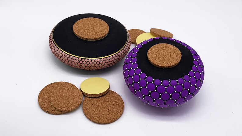 May include: Two round wooden boxes with cork lids. The box on the left is painted pink with black and gold accents. The box on the right is painted purple with black and white accents. Both boxes are decorated with a pattern of small circles.