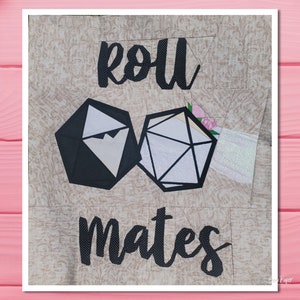 Puede incluir: Un edredón beige con las palabras "roll mates" en escritura negra. Se presentan dos formas geométricas de dados, una negra y otra blanca, con un acento floral. El edredón tiene un diseño estampado.