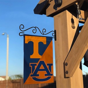Puede incluir: Una bandera azul marino y naranja con las letras "T" y "AU" se muestra en un soporte de metal negro. La bandera cuelga de una estructura de madera, con una bombilla visible en el fondo.