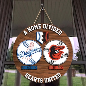 May include: A round wooden sign with the text "A HOME DIVIDED HEARTS UNITED". The sign features baseball logos for the Dodgers and Orioles. The Dodgers logo is blue and white, and the Orioles logo is black and orange.