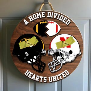 Puede incluir: Letrero redondo de madera con un balón de fútbol dividido y dos cascos. El texto dice "A HOME DIVIDED HEARTS UNITED". El balón está dividido, con negro y dorado por un lado, y rojo y blanco por el otro.