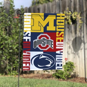 Puede incluir: Una bandera de jardín rectangular con los logotipos de la Universidad de Michigan, la Universidad Estatal de Ohio y la Universidad Estatal de Pensilvania. La bandera tiene un fondo azul, rojo y blanco con las palabras "HOUSE DIVIDED" impresas verticalmente.