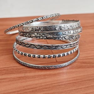 May include: A stack of silver bangles with intricate designs. The bangles feature floral, dotted, and patterned details. The bangles are displayed on a wooden surface.