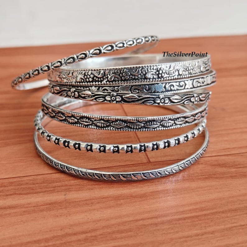 May include: A stack of silver bangles with intricate designs. The bangles feature floral, dotted, and patterned details. The bracelets are displayed on a wooden surface.