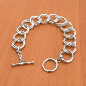 Op de afbeelding: Een zilverkleurige kettingarmband bestaande uit in elkaar grijpende dubbele ronde schakels. De armband is voorzien van een knevelsluiting bestaande uit een T-balk en een enkele ronde ring, wat zorgt voor een veilige sluiting van het sieraad.