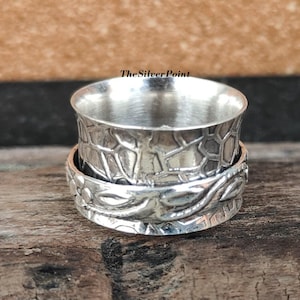 May include: A silver spinning ring with a textured design. The ring has a floral pattern on the bottom band and a geometric pattern on the top band.