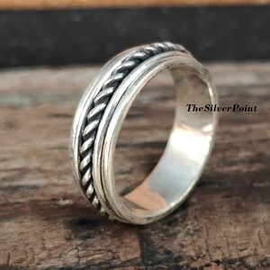 Könnte beinhalten: Ein silberner Ring mit einem geflochtenen Design. Der Ring liegt auf einer Holzoberfläche. TheSilverPoint ist auf der Oberfläche geschrieben.