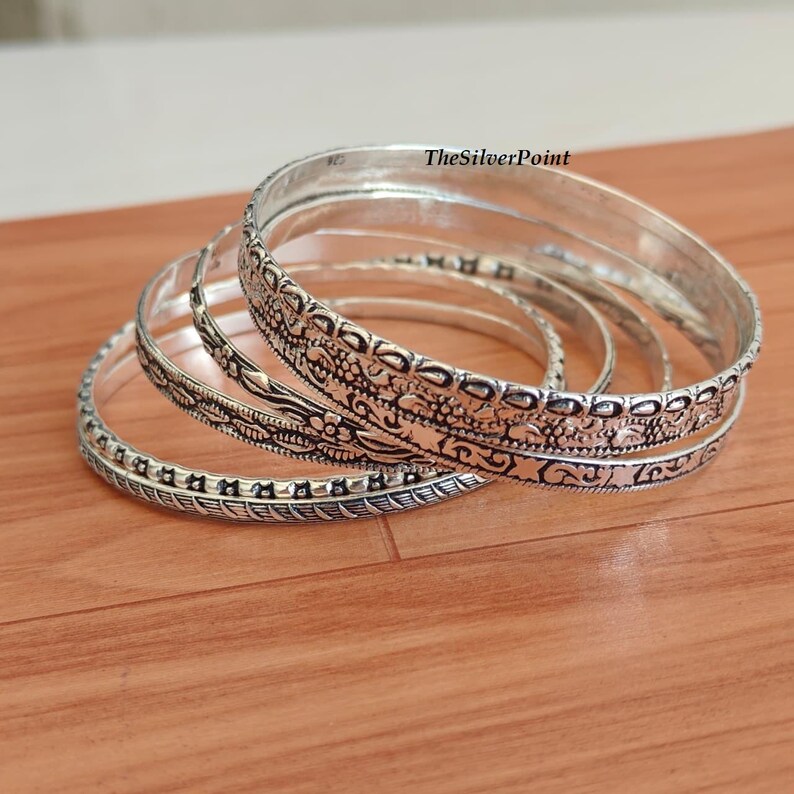 May include: Several silver bangles with intricate floral and geometric patterns are arranged on a wooden surface. The bangles vary in design, with some featuring detailed engravings and others with simpler, textured bands. TheSilverPoint is visible on the top right.