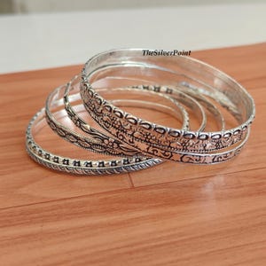 May include: A collection of silver bangles with intricate floral and geometric patterns. The bangles are stacked on a wooden surface, showcasing their detailed designs and reflective qualities. TheSilverPoint is visible on the top right.