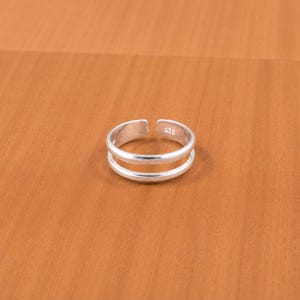 Puede incluir: Un anillo abierto de plata con un diseño de doble línea. La banda de metal tiene un acabado pulido e incluye un pequeño sello 925 claramente visible en la superficie interior, lo que indica la composición de plata esterlina de esta joya.