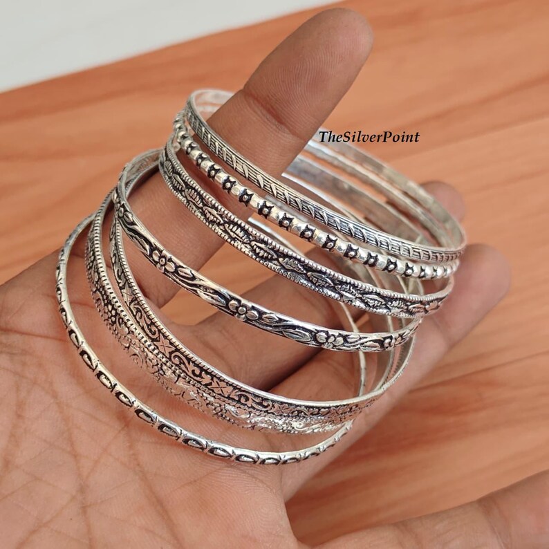 May include: A collection of silver bangles with intricate etched designs. The bangles are held in a hand, showcasing the various patterns, including floral and geometric motifs. The text "TheSilverPoint" is visible.
