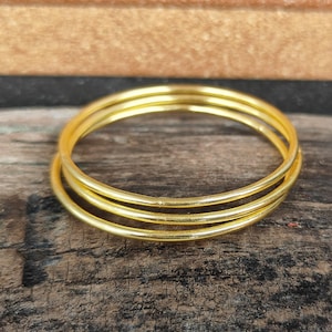 May include: Three gold-colored bangle bracelets stacked on top of each other on a wooden surface.