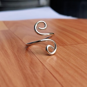May include: A silver-colored metal ring featuring an open-ended design with two symmetrical spiral-shaped terminals. The band is smooth and polished, curving into distinct coils at each end, creating a continuous, fluid, and reflective metallic form.
