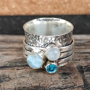 Peut inclure: Une bague tournante en argent avec un motif floral et trois pierres. Les pierres sont bleu clair, pierre de lune blanche et opale blanche.
