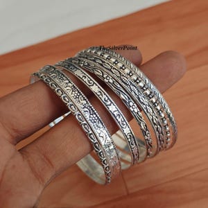May include: A collection of silver bangles with intricate floral and geometric patterns. The bangles vary in design, with some featuring beaded accents. The jewelry is displayed on a wooden surface, showcasing the detailed craftsmanship of each piece.