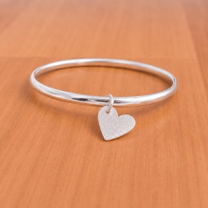 May include: A polished silver bangle bracelet featuring a single heart-shaped charm. The bangle has a smooth, reflective surface, while the heart charm displays a textured, matte finish. The charm is attached to the bangle by a small silver ring.