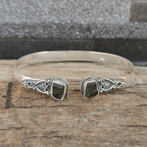 May include: Silver bangle bracelet with two dark green gemstone accents. The bracelet has a delicate, ornate design with a heart-shaped motif.